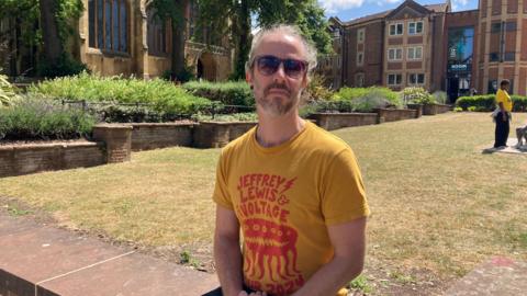 A man with wavy white hair sits on a low wall surrounding a grassy area. He is wearing a yellow T-shirt with some red writing on and sunglasses. There is a church and some buildings surrounding the green behind him.