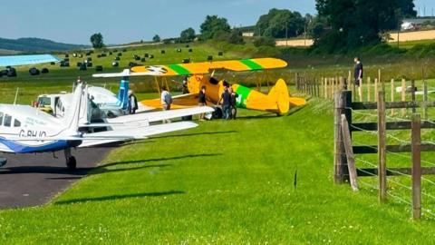 A yellow and green plane parked on a grass verge next to several small white and blue planes.