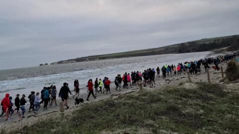 Line of people running on the sand along a beach with grass and sand to the right and the sea to the left.