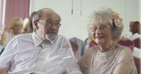 Man in shirt and tie in glasses on the right. He's holding some flowers in his hand. He's golding the hand of the women on the right. She has glasses and curly hair. She has a flower in hair and is smiling.
