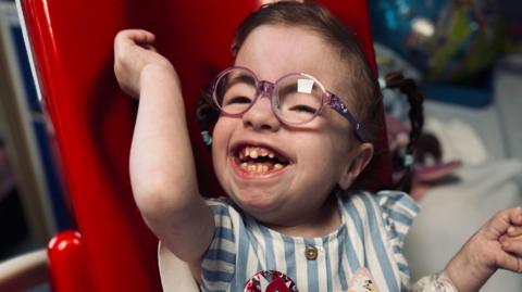 Ella has a big smile as she sits in a high-chair. She is wearing purple rimmed glasses and a blue and white top.