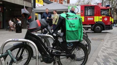 An e-bike is parked in between bike railings with a large green Uber Eats bag attacked to the back of it. In the background, a red London Fire Brigade fire engine is parked behind a grey marquee.