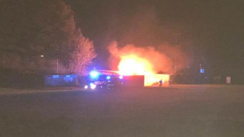 A large fire in a school playing field. A large tree is in the field, with another to the side. There are firefighters trying to put the fire out and a fire engine parked up too. 