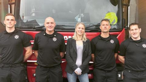 A woman stood with two men on either side of her. They are all stood in front of  a fire engine.