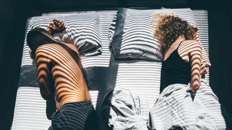 A couple sleeping in the same bed. They are both turning away from each other and are far apart. The sun is shining on the bed and the shadow of the blinds is on them.