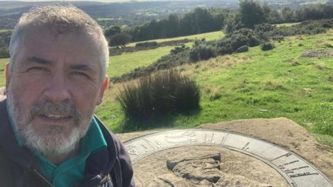 Nick Burton stands at the memorial to the late fell walker and writer Alfred Wainwright. The piece of stone has a picture of Wainwright carved into it. Nick stands on the left, he has short grey hair, a grey beard and wears a black coat with a teal polo shirt under it.