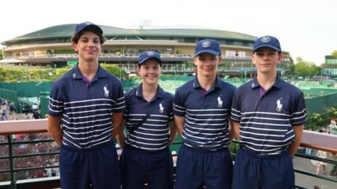 A group of four children wearing blue polo shirts with white hoops on the bottom. They have their arms behind their back and are smiling. They are stood in front of a stadium, with tennis courts also behind them.