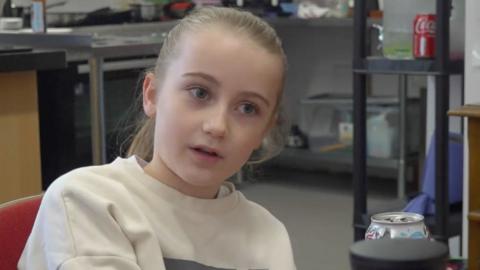 Primary school pupil, she is sat in a Cafe. She has her hair tied back in a ponytail and is wearing a white jumper.