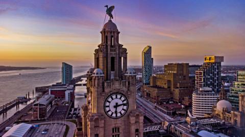 The sun setting behind the Royal Liver Building in Liverpool, with the cityscape and mouth of the River Mersey seen in the background. 