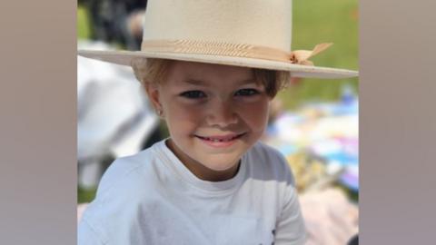 Ruby is wearing an oversized straw hat and is smiling widely at the camera. She is also wearing a white T-shirt and is sitting on a picnic mat.