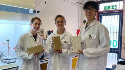 Three members of the earth4Earth company, wearing white lab coats, in their research facility at Sheffield Technology Parks. They each hold one of the "carbon absorbing" bricks. Two of the bricks are beige in colour and the other brick is an off-white colour.