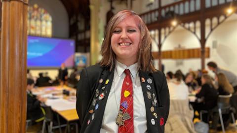 Lexi smiles at the camera. Behind her students are sitting and talking to each other in a school hall. Lexi is wearing a black blazer with lots of badges pinned on it and on her red tie. Her hair is dyed different colours and is brushed to one side.