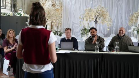 Three people are sitting behind a black table. They are looking towards a woman who is standing with their back towards the camera. She is wearing a red sweater vest and a white shirt.