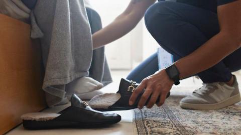 A nurse helps a person with their shoes at home. The close-up photograph shows a nurse crouching on the floor. The person receiving care is sitting on the bed wearing socks. Daylight is shining through the window and there is a patterned rug by the bed.