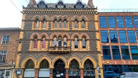 A historic brick building with decorative arches and patterned stonework stands along a town street, with bicycles parked outside and a modern building beside it.