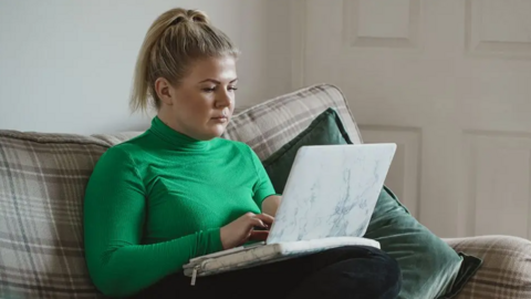A young woman is sitting on a patterned settee and using a laptop. She is wearing a green jumper and has blonde hair tied back.