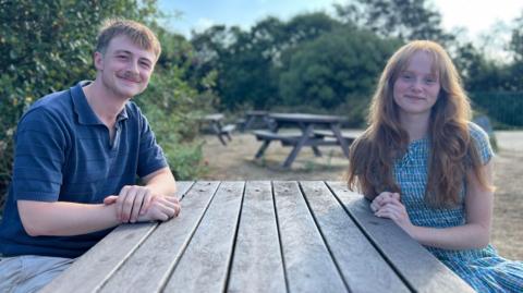 Bill Nicholson is wearing a blue top and sitting at a picnic table opposite Amber Thomson who is wearing a blue dress.