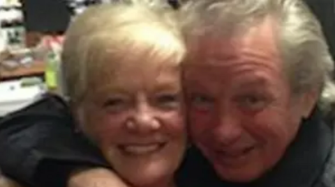 Photograph of Sheila Jackson, who has blonde hair, and Eric Greener, who has grey hair. Mr Greener has his arm around Ms Jackson's shoulder and they are both smiling for the camera in the close-cropped shot. 