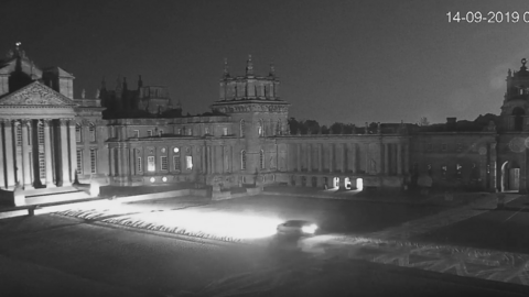 A night CCTV image of Blenheim Palace courtyard showing a car driving through it with headlights on. The ground leading up to the palace is overed in large Union flags