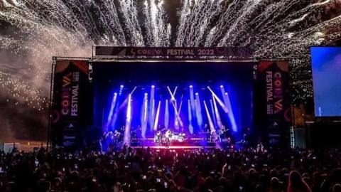 A band playing at a big stage at Godiva Festival with blue and purple lights as well as fireworks. A large audience of people is in front of the stage.