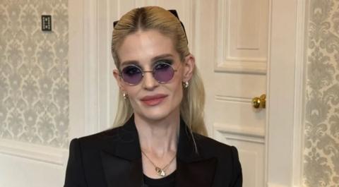 Kelly pictured slightly smiling while wearing purple lens circular glasses and a black blazer. She has blonde hair scraped back away from her face.