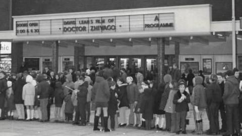 A black and white photograph showing a long line of people queuing to get into the old Odeon cinema when it was open.