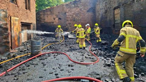 Firefighters in protective equipment tackle a blaze at a heavily fire-damaged industrial building.
