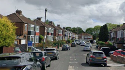 Hillcrest Avenue is a street with two-storey brown brick semi-detached houses. There are parked cars on each side of the road. Several trees can be seen in the distance and to the sides.