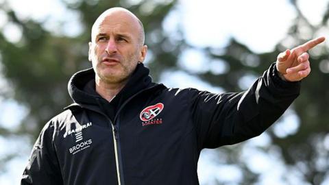Gavin Cattle is pointing to somewhere on the pitch. He is bald and is wearing a waterproof coat with the Cornish Pirates badge on it.