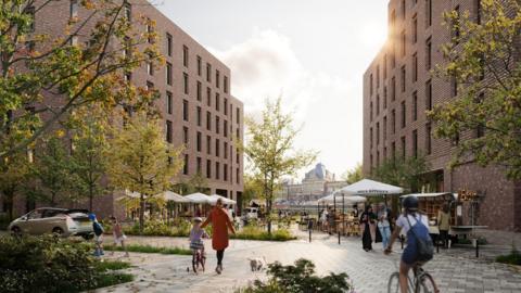 A pedestrian-friendly urban courtyard framed by mid-rise brick buildings, with trees in autumn foliage. People walk, cycle, and sit at outdoor cafe tables under white umbrellas labelled “Soul Kitchen.” In the background Norwich railway station is visible.