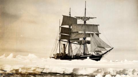 Black and white image of the Terra Nova ship sailing in Antarctica. The ship is wooden, it's sails are open. It is moving across ice-covered ocean. 