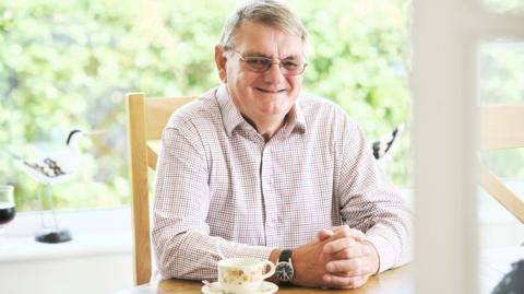 Photo of David oar smiling and sitting at a table. He is a middle-aged man with glasses and a lightly coloured checked shirt.
There is a cup and saucer on the table.