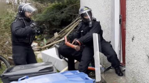 Two police officers breaking into a run-down property with rubbish covering the back garden. One officer uses a battering ram and is smashing in the property door. Both are wearing protective head gear and bullet proof vests.