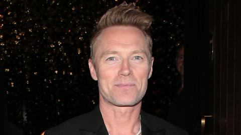 Roanan Keating has light brown hair pushed up high on his head. He is wearing a dark black jacket and the background behind him is dark.