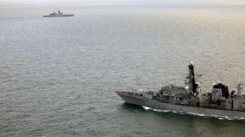 HMS Somerset sails in the sea as RFS Soobrazitelny also sails in the sea in the distance. The water looks calm. HMS Somerset has F82 written on its side.