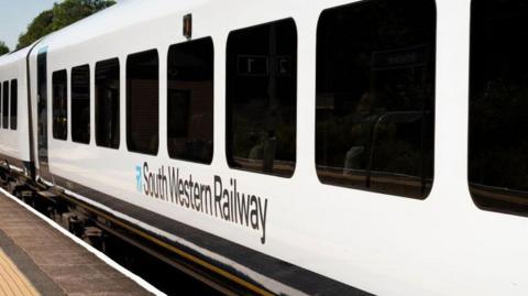 A white train with 'South Western Railway' on the side in black lettering is at a train platform.