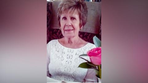 Maureen Sanderson, wears a white top and is smiling slightly. A pink flower is in the fore front of the right hand side of the picture.