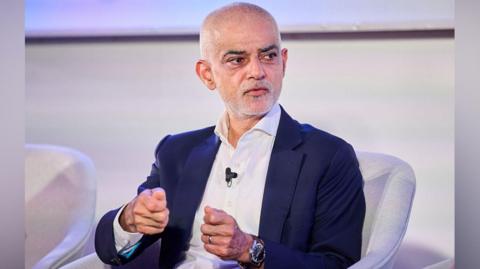 Sir Sadiq Khan sitting on a cream-coloured chair with a similar coloured background. He is wearing a radio mic and a dark blue suit jacket with a white shirt. 