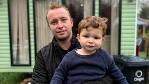 Eoin McGovern is standing outside a green mobile home and is holding his young son