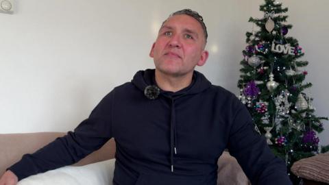 A man in a black jacket and jeans, is sitting with his white dog on a sofa. 