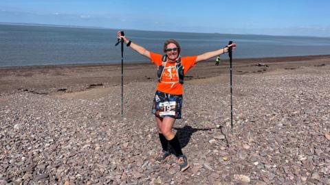 Channel Events runner on the beach with their arms outstretched holding walking poles, smiling at the camera