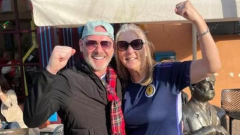 Jim and Diane Lees have their arms around each other. Jim wears a tartan scarf, black jacket and backwards light blue cap. He has short white hair. Diane wears a Scotland strip and has long blonde hair and both raise their fists in the air.
