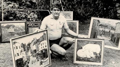 A man crouching down in a garden with dark, curly hair wearing a white short sleeved shirt and dark coloured shorts holding two pictures he painted. One on the right of two pigs and the other on the left of Park Street in Bristol. Six other paintings are behind him in front of a hedge.