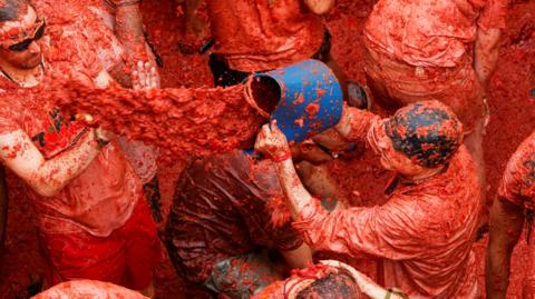 Captured from above, a group is covered in tomato juice and a man - on the right - is throwing a bucket of the juice onto someone else