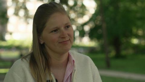 Charlotte, who has long light-brown hair and is wearing a white cardigan and pink and white striped shirt, is sitting in a park 