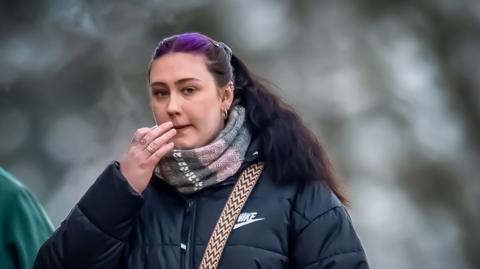 Rhiannon Evans arriving outside court. She has pulled back long black hair with a purple streak around her top part and is wearing a puffy black winter coat.