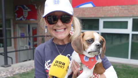 A woman wearing a white England cap and England earrings holds her dog to the camera.