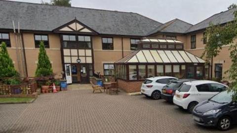 Hallgarth Care Home is a beige-brick two-storey building with a grey roof. It has a small seating area outside and a several car parking spaces.