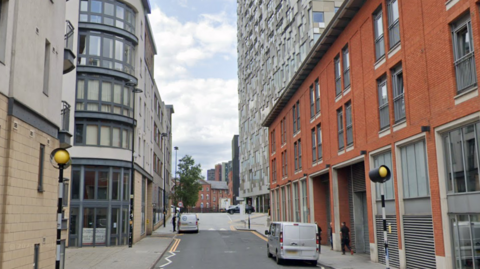 A road outside the Mailbox which has a zebra crossing, and flats on either side of the road. There are two silver vans parked on the road.