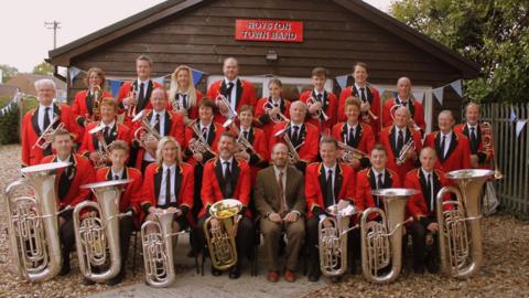 Photograph of band wearing red blazers, black ties and holding their brass instruments.